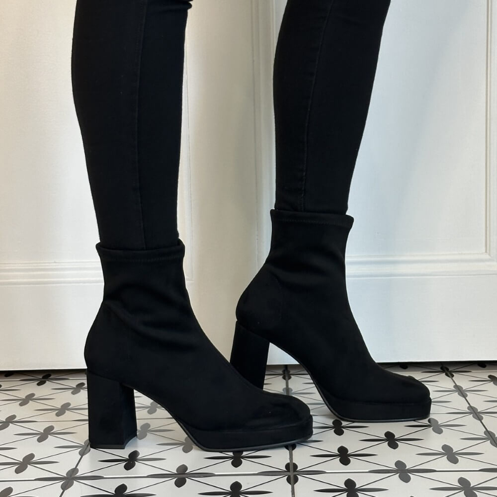 Miss Elastic Black Ankle sock boots with chunky block heels, shown on a model wearing black skinny pants against a white door and patterned tile floor.