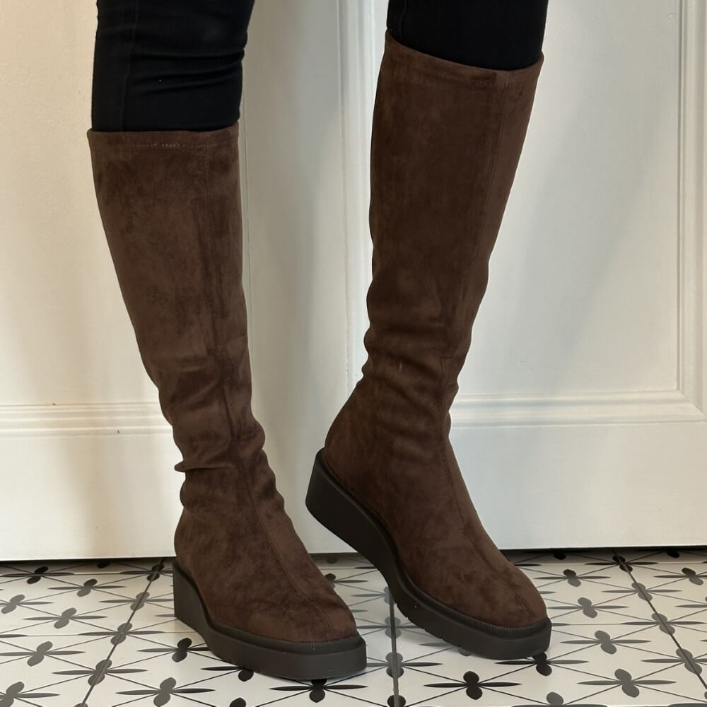 A pair of Miss Elastic knee-high brown suede boots with chunky black platform soles, shown on a model wearing black leggings in front of a white panelled door.