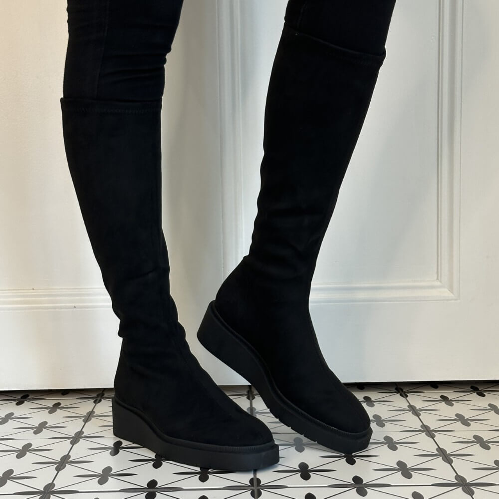 A pair of Miss Elastic black over-the-knee platform boots with a matte suede-like finish, shown on a model wearing black skinny pants against a white panelled wall and patterned tile floor.