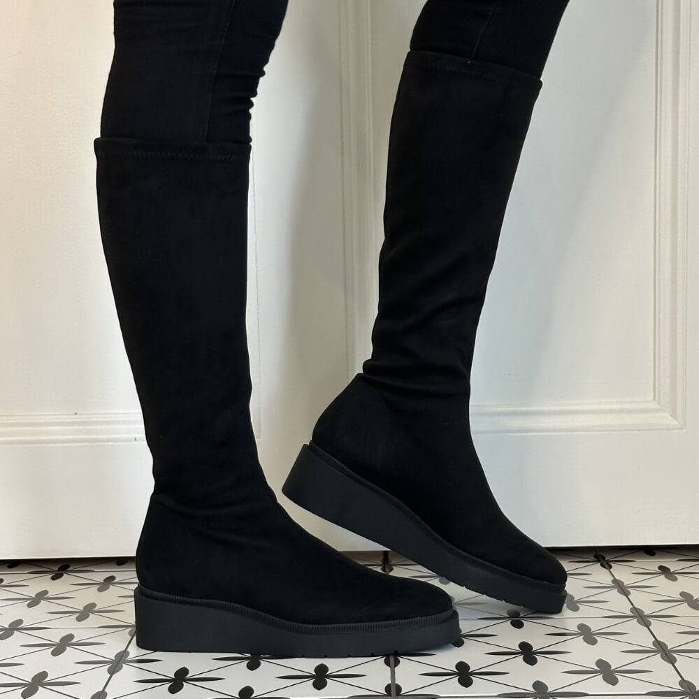 A pair of Miss Elastic black over-the-knee platform boots with a matte suede-like finish, shown on a model wearing black skinny pants against a white panelled wall and patterned tile floor.