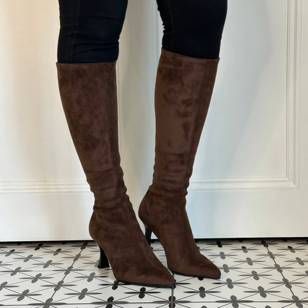 A pair of Miss Elastic knee-high brown suede boots with a kitten heel, worn with black leggings, standing on a black and white patterned tile floor in front of a white panelled wall.