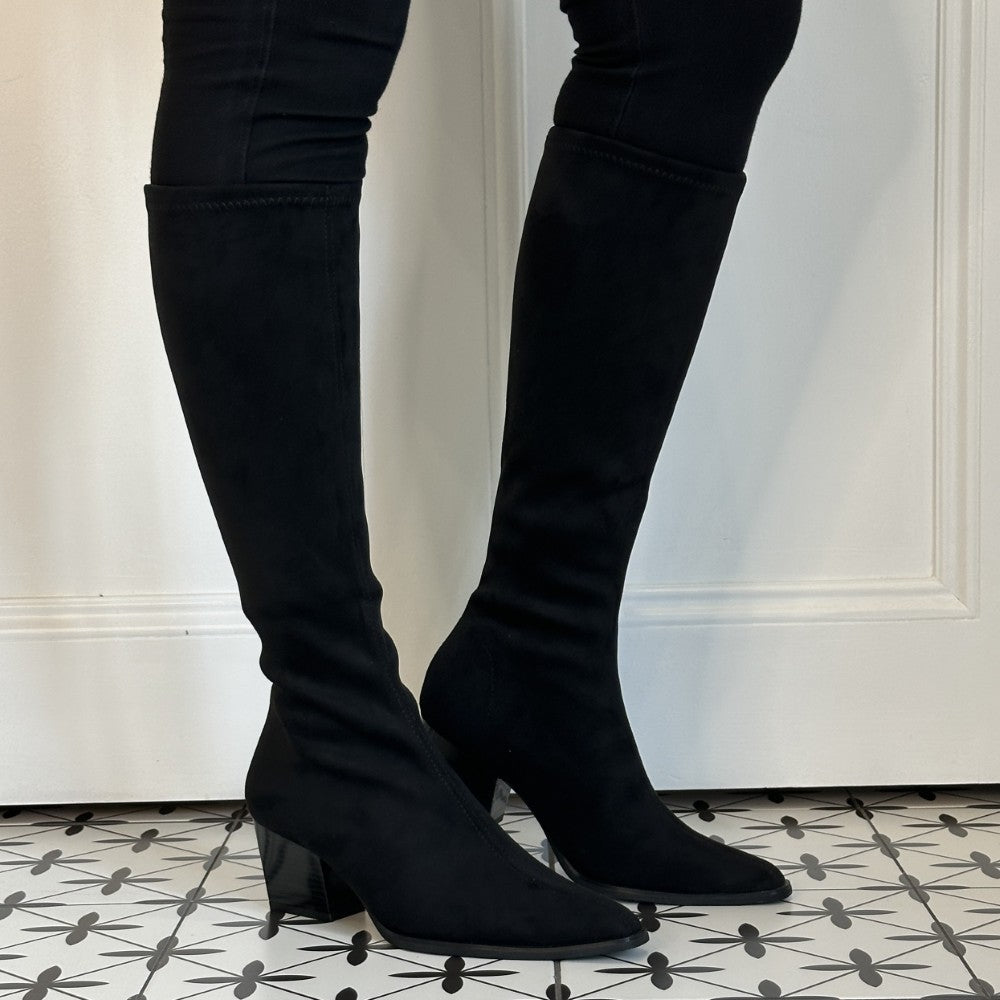 Miss Elastic Black suede knee high boots with a block heel, shown on a model wearing black skinny jeans against a white panelled wall and black-and-white patterned tile floor.