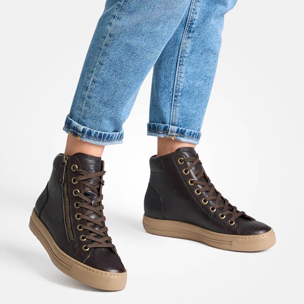 A person wearing Paul Green dark brown leather high-top lace-up sneakers with side zippers, brown laces, and tan soles, paired with cuffed blue jeans against a light gray studio background.