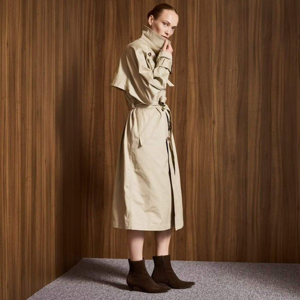 Woman wearing Brown Boots a beige trench coat against a wooden paneled wall.