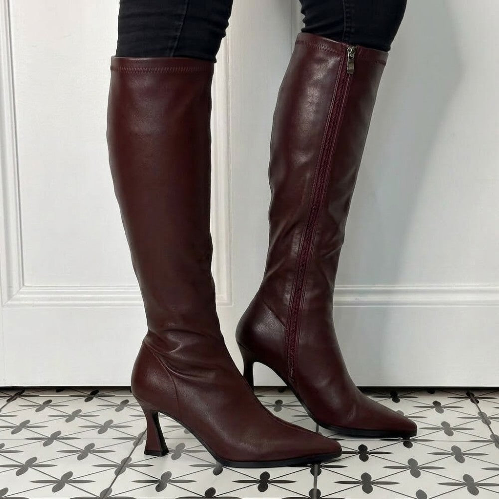 A pair of Pedro Anton Wine knee-high leather boots with pointed toes and kitten heels, shown on a model's legs against a white panelled door and patterned tile floor.
