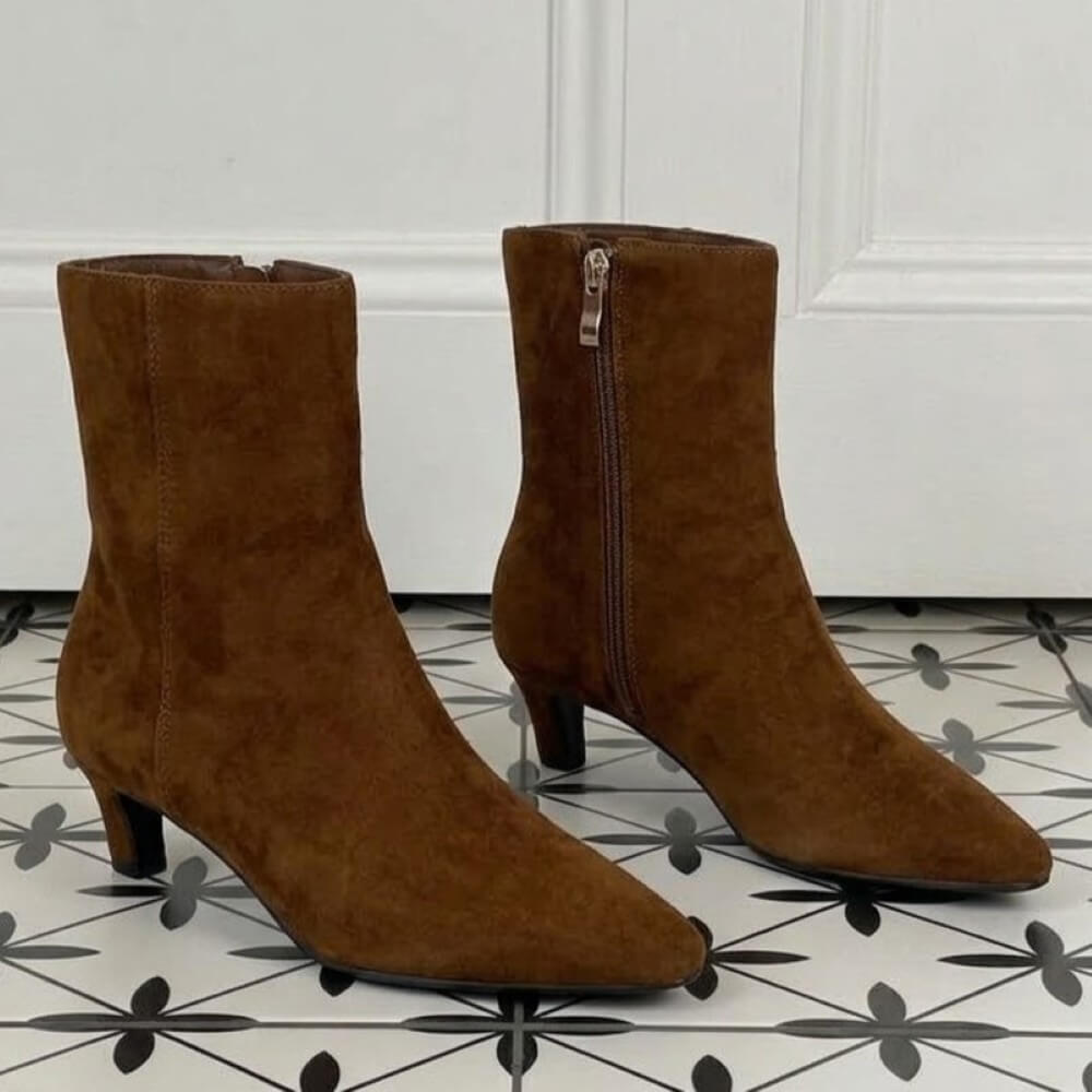 A pair of Pedro Anton Tan suede ankle boots with pointed toes and low block heels, standing on a black and white patterned tile floor in front of a white panelled wall.