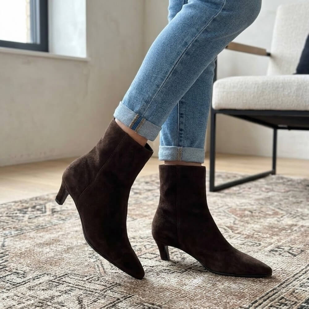 A person wearing Pedro Anton dark brown suede ankle boots with kitten heels and pointed toes, paired with light blue cuffed jeans, standing on a patterned rug in a modern room.