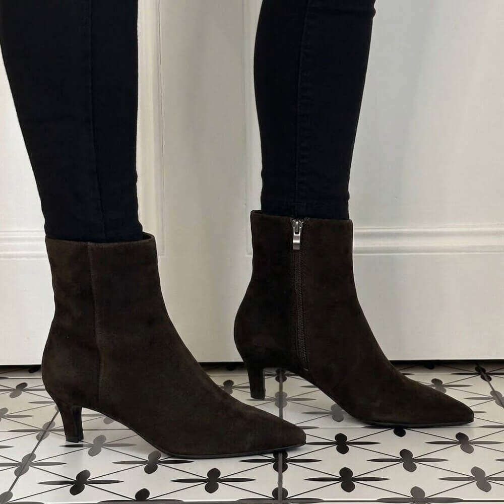 A pair of Pedro Anton dark brown suede ankle boots with kitten heels and side zippers, worn with black skinny jeans on a black and white patterned tile floor.
