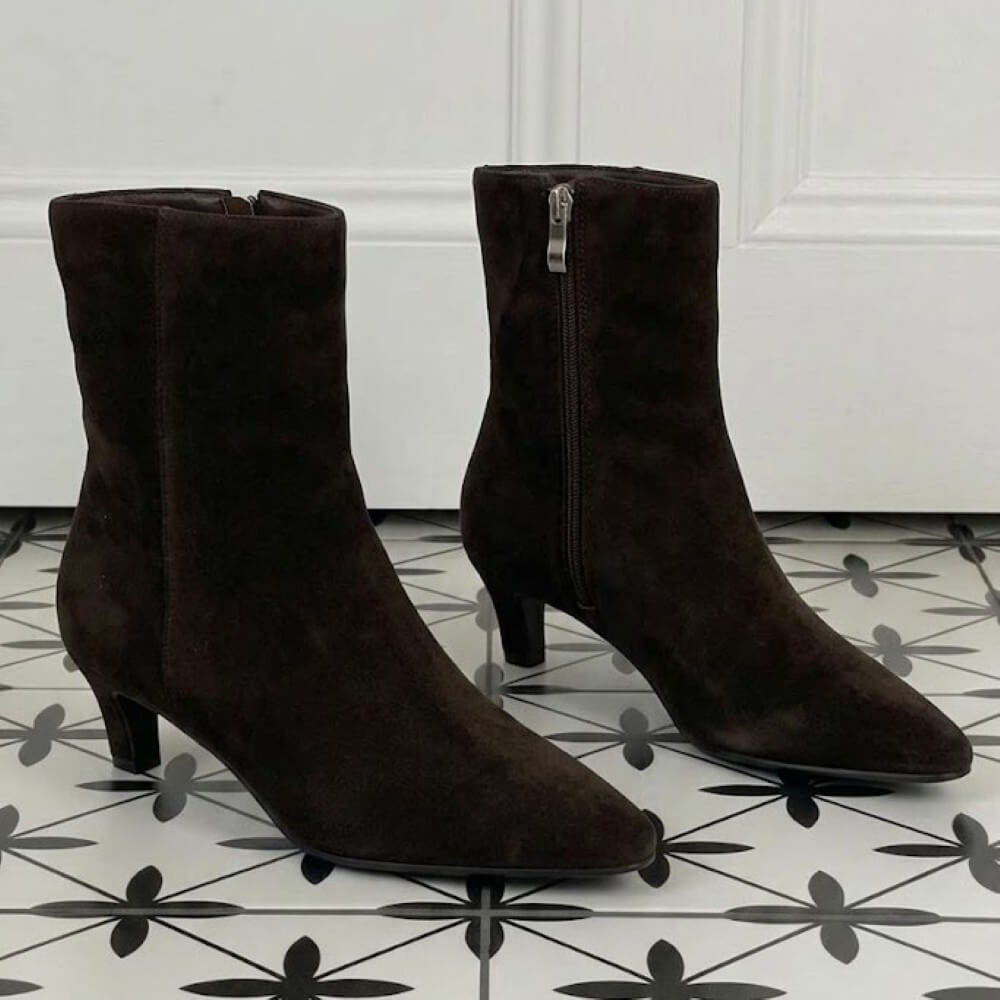 A pair of dark brown suede ankle boots with kitten heels and side zippers, displayed on a black and white patterned tile floor in front of a white paneled door.