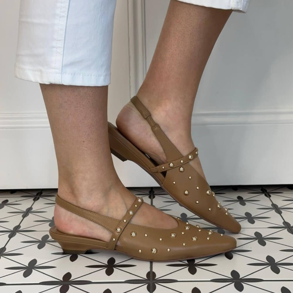 Pedro Anton Brown leather studded slingback flats worn with white pants on a patterned floor.