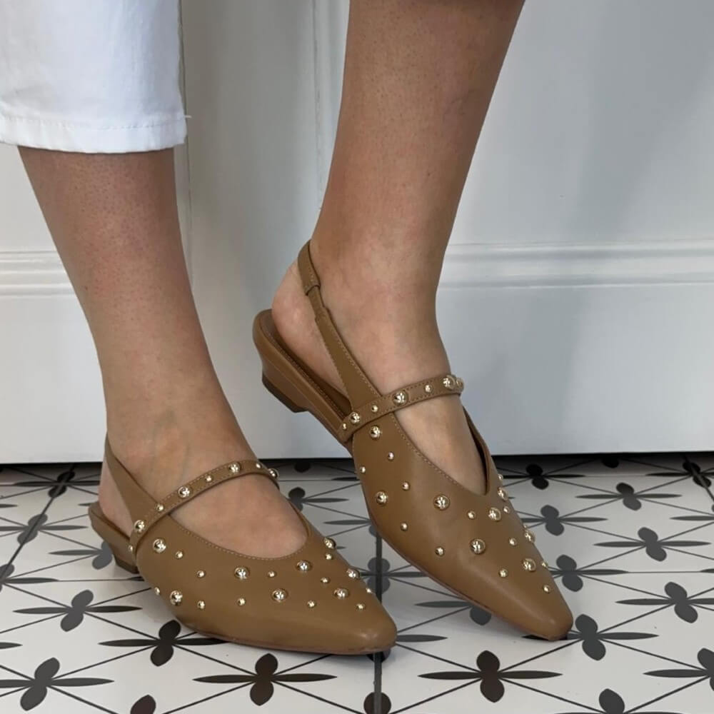Pedro Anton Brown leather studded slingback flats worn with white pants on a patterned floor.
