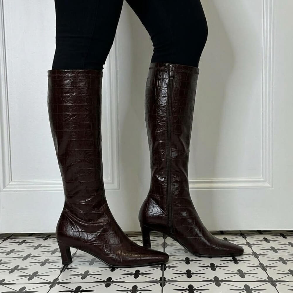 A person wearing Pedro Anton knee-high dark brown crocodile-embossed leather boots with kitten heels, standing on a black and white patterned tile floor in front of a white panelled wall.