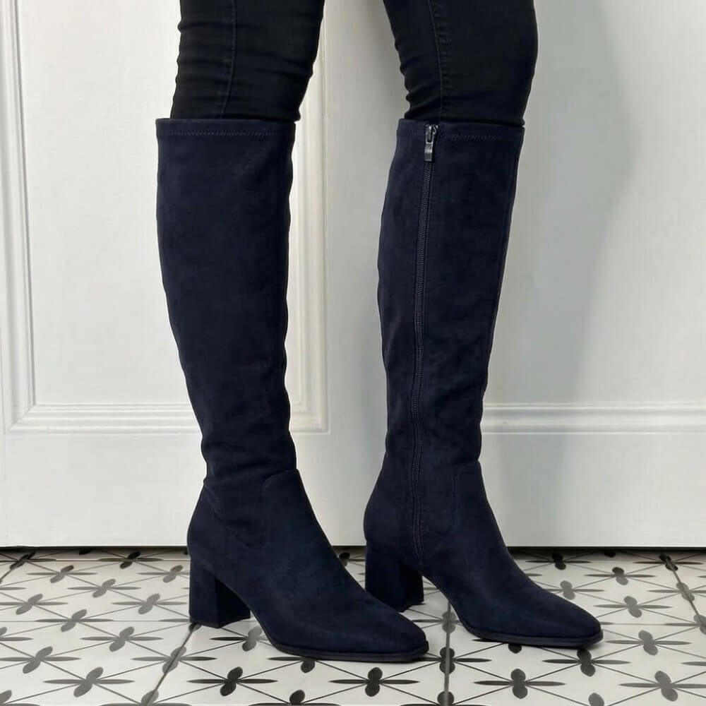 A pair of Pedro Anton dark navy blue suede knee-high boots with block heels, worn with black skinny jeans, standing on a black and white patterned tile floor in front of a white panelled wall.