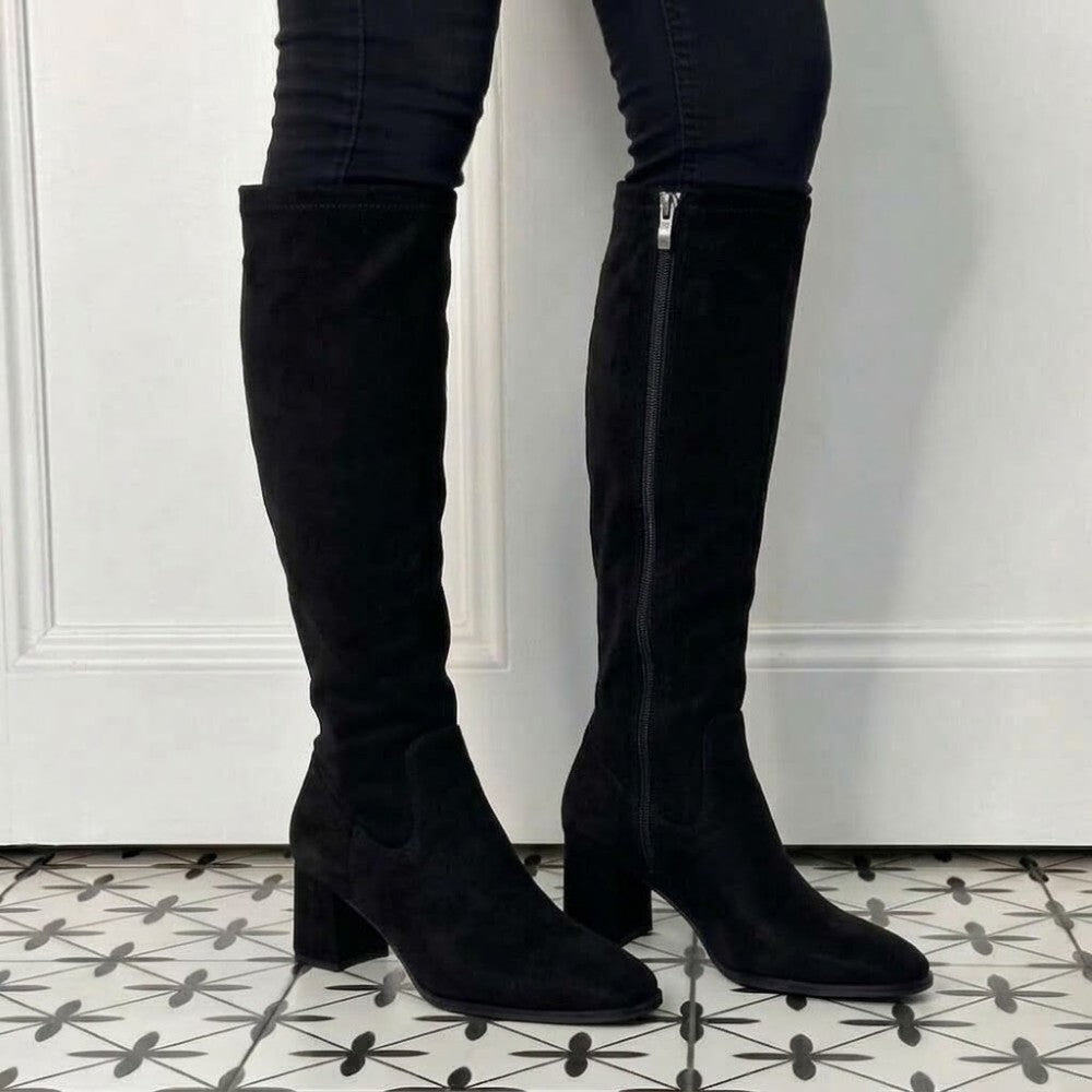 A pair of Pedro Anton black suede knee-high boots with block heels and side zippers, shown on a model wearing black jeans against a white Pedro-Anton-Yasmine-Knee-High-Navy-Suede-Boot-a door and patterned tile floor.