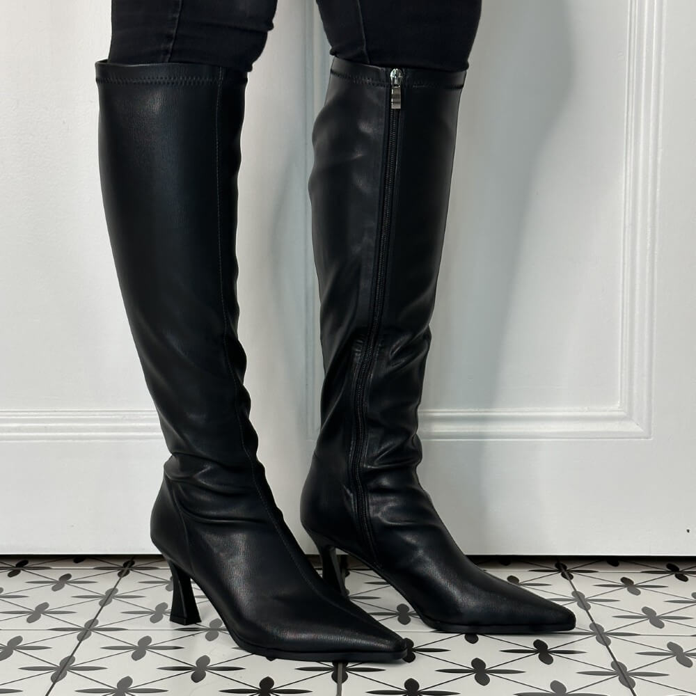 A pair of Pedro Anton black leather knee-high boots with pointed toes and kitten heels, shown worn with black pants against a white panelled wall and patterned tile floor.