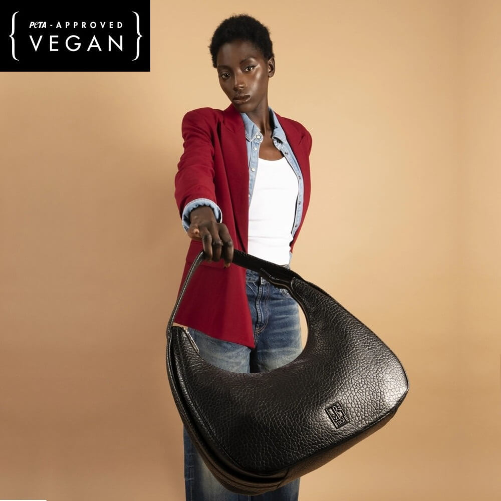 Woman holding a black leather handbag against a beige background with 'PETA APPROVED VEGAN' label.