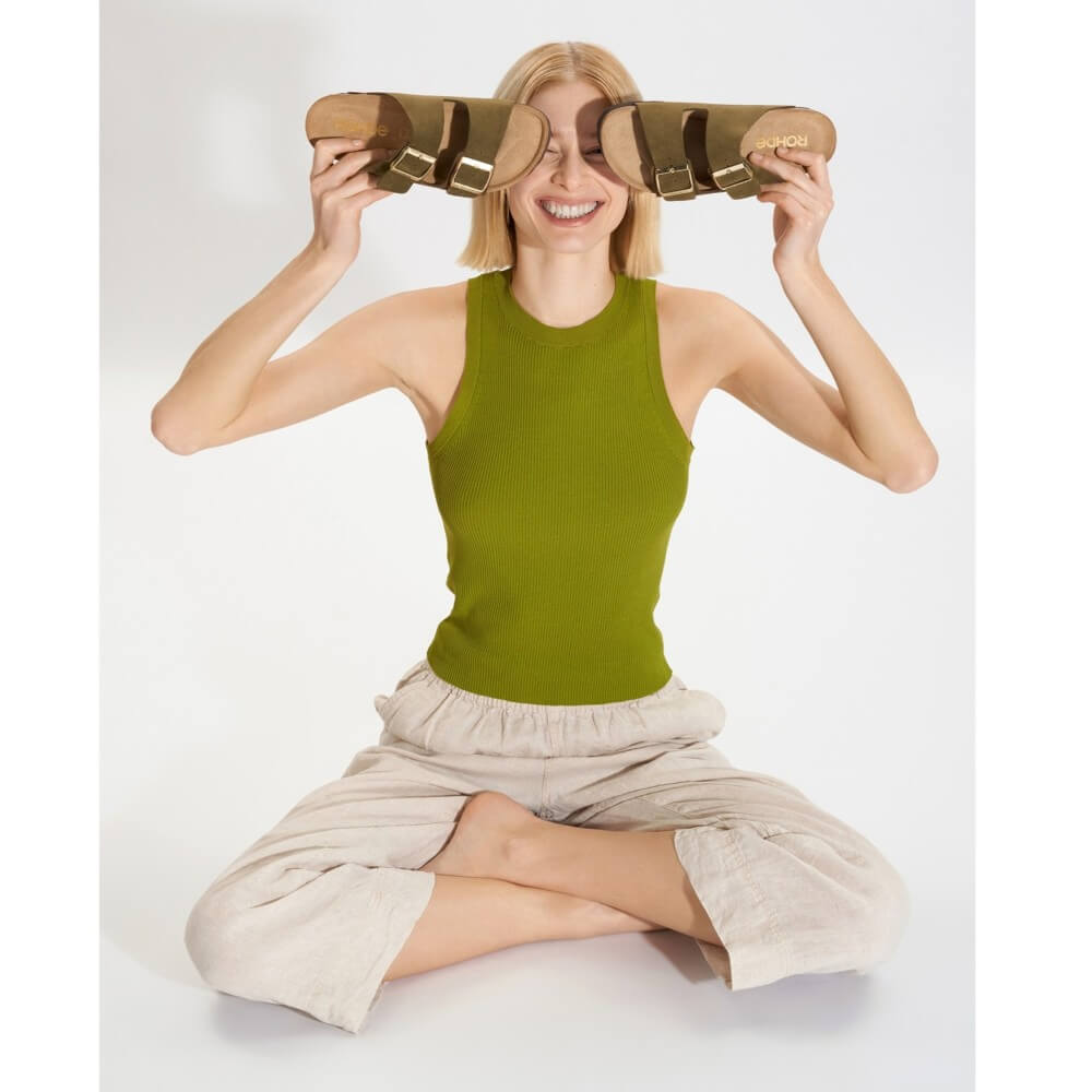 Person wearing a green sleeveless top and beige pants sitting cross-legged, holding Rohde sandals above their face on a white background.