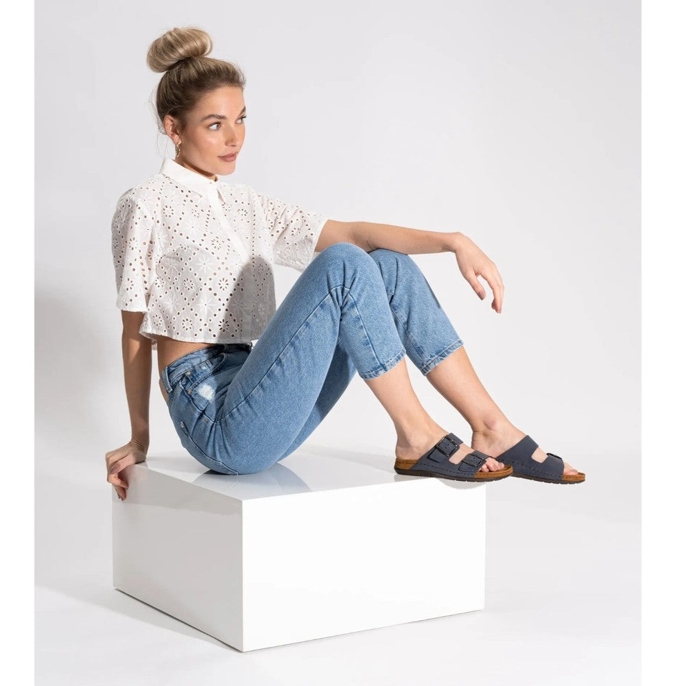 Woman sitting on a white cube wearing a white blouse, blue jeans, and navy Rohde sandals.