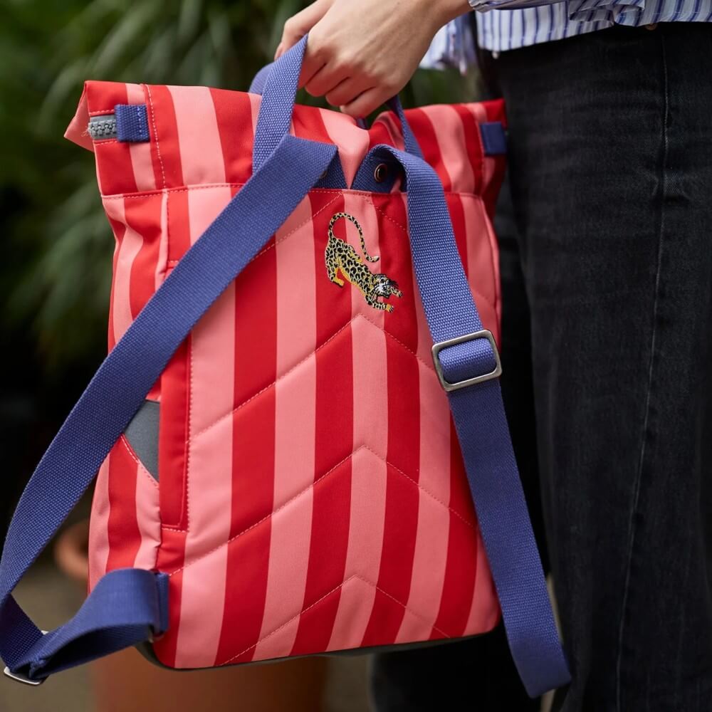 Roka Red and pink striped bag with blue straps held by a person, against a blurred green background.