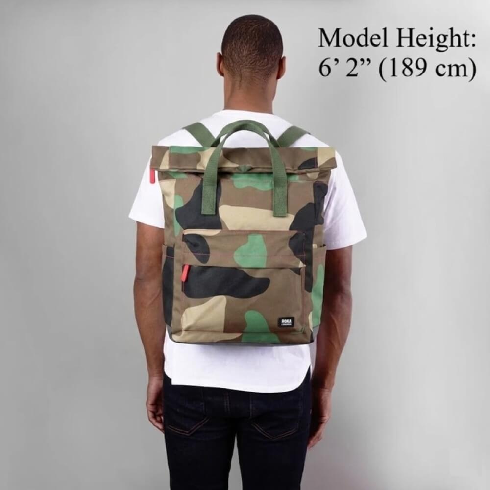 Person wearing a camouflage Roka backpack with model height information on a grey background