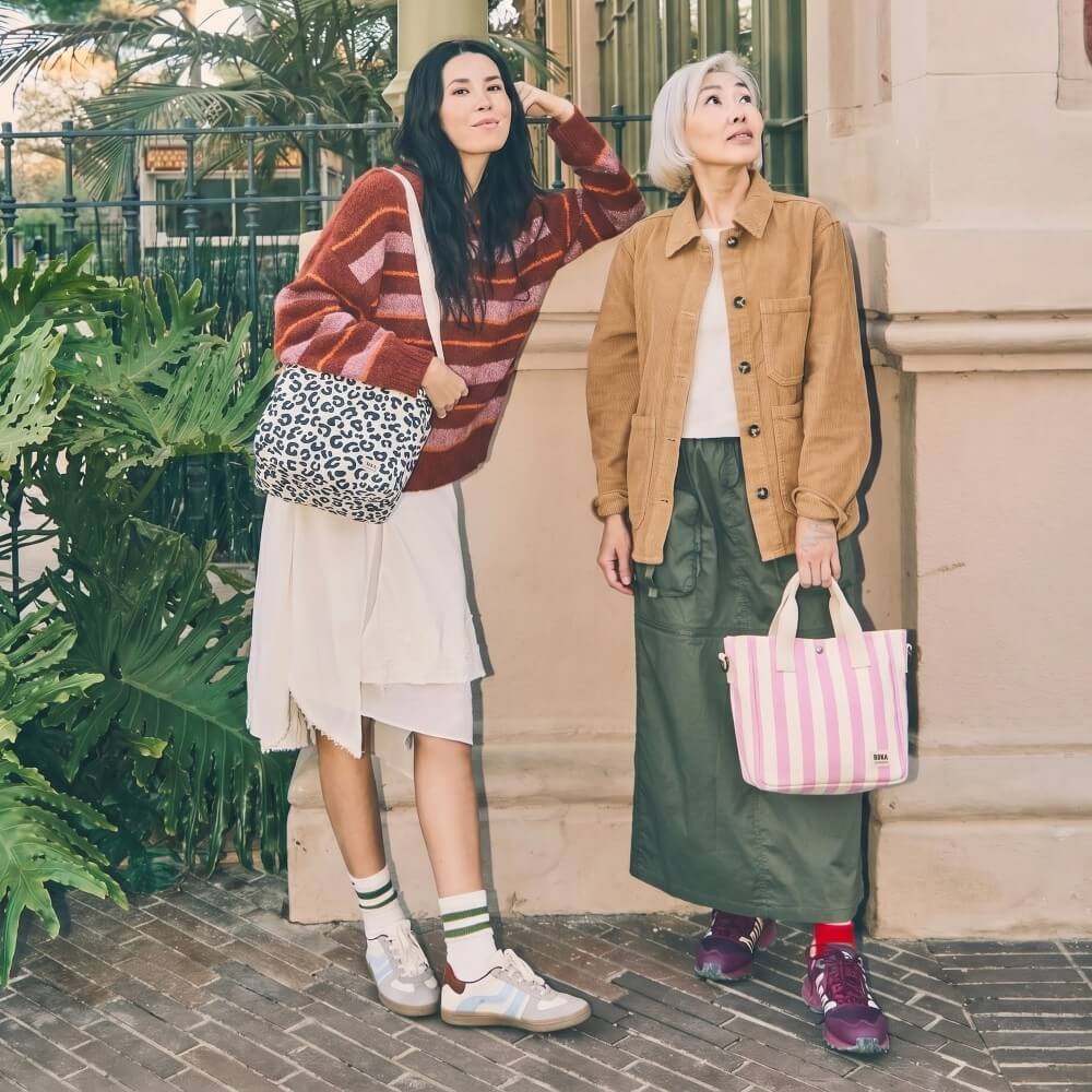 Two women standing outdoors, both holding Roka lunch bags, against a building and plant background.