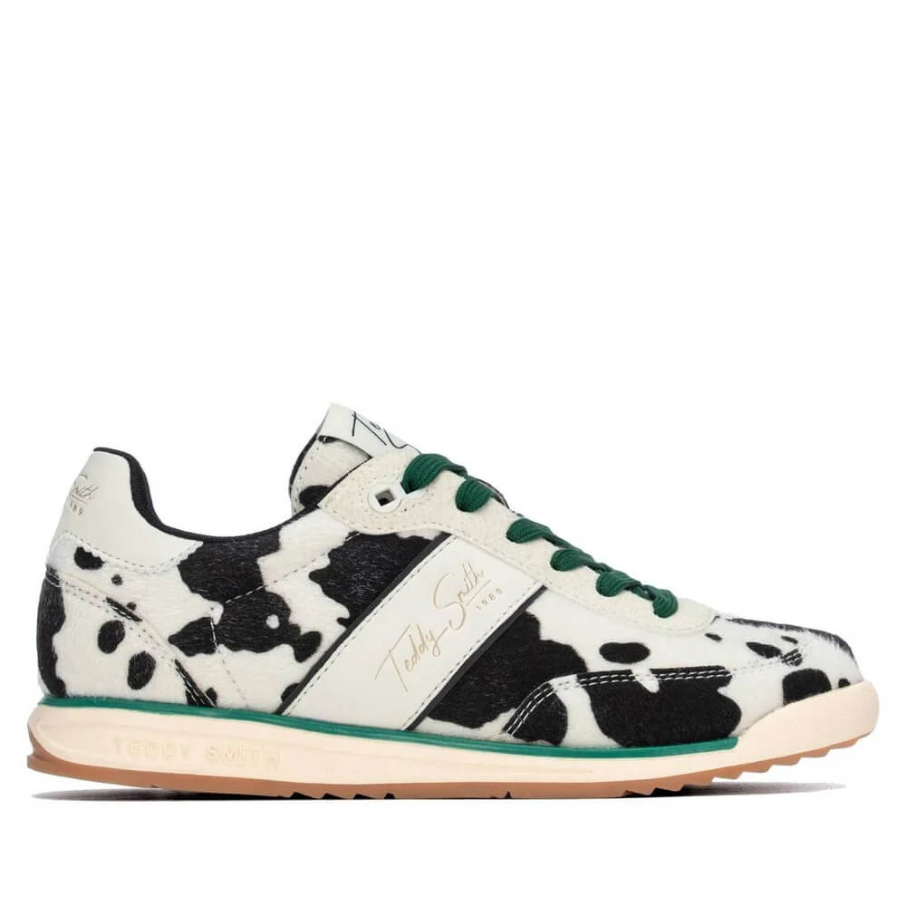 A low-top sneaker by Teddy Smith featuring a black and white cow print pattern, green laces, a white side stripe with gold script branding, and a beige midsole with a gum outsole.