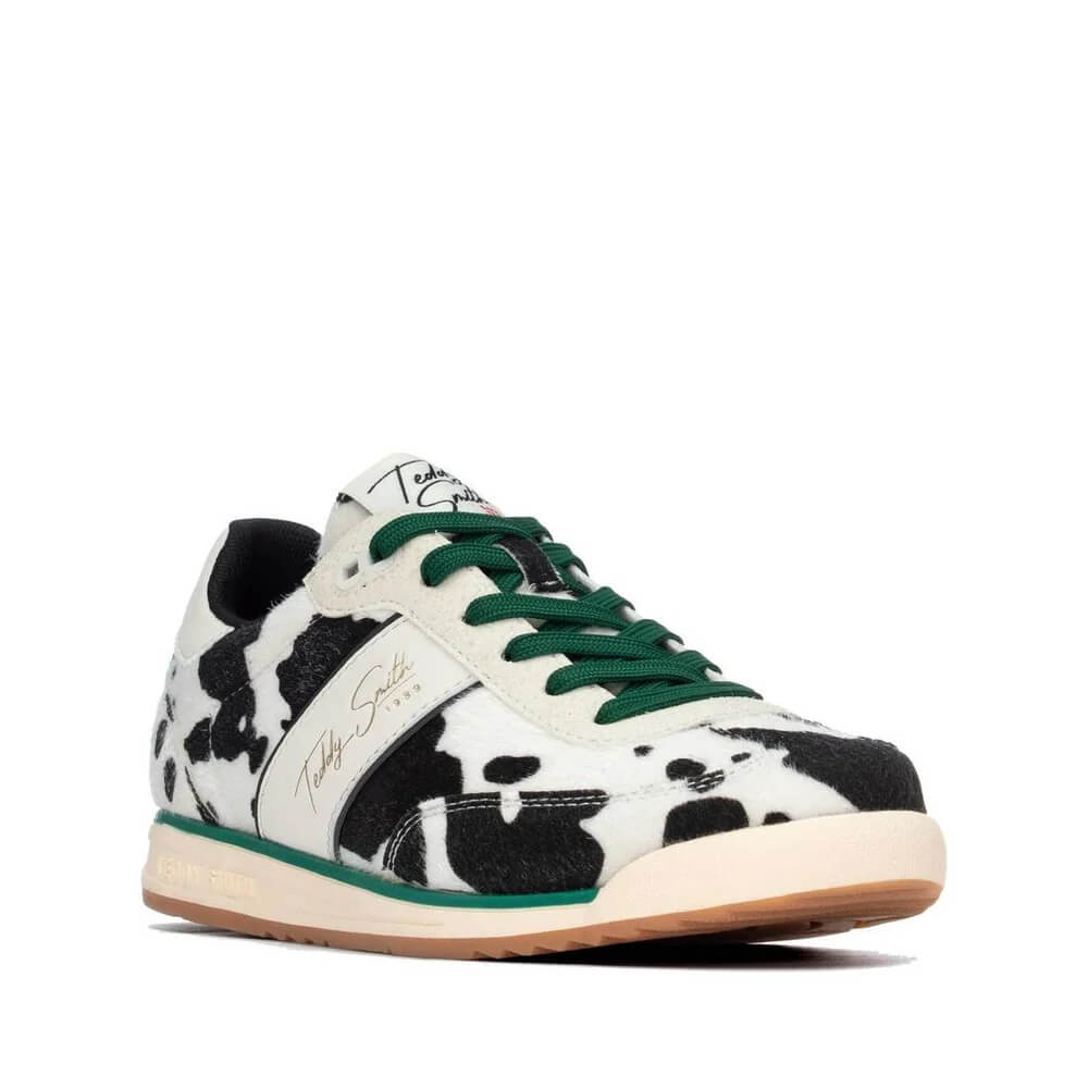 A low-top sneaker by Teddy Smith featuring a black and white cow print pattern, green laces, a white side stripe with gold script branding, and a beige midsole with a gum outsole.