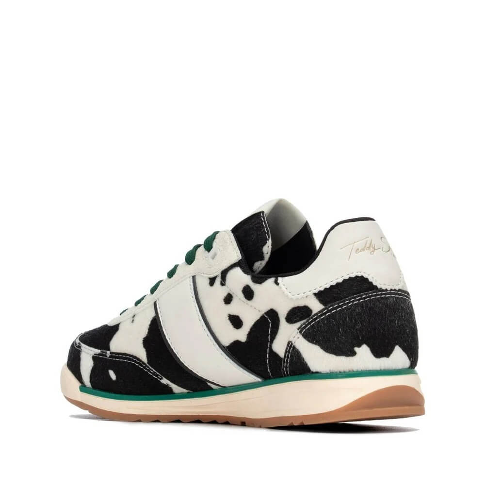 A low-top sneaker by Teddy Smith featuring a black and white cow print pattern, green laces, a white side stripe with gold script branding, and a beige midsole with a gum outsole.