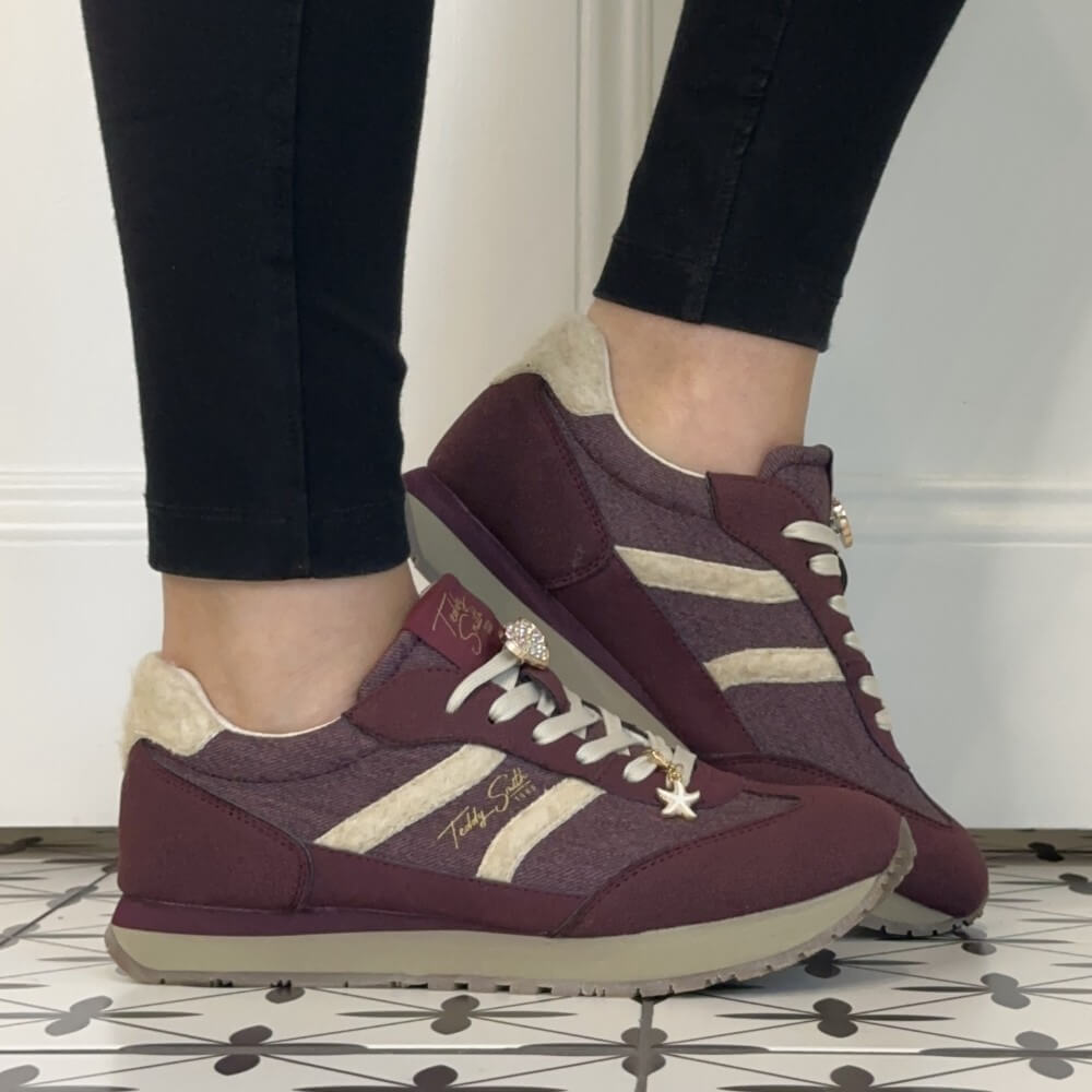 A person wearing Teddy Smith burgundy and beige low-top sneakers with black pants, standing on a patterned tile floor against a white wall.