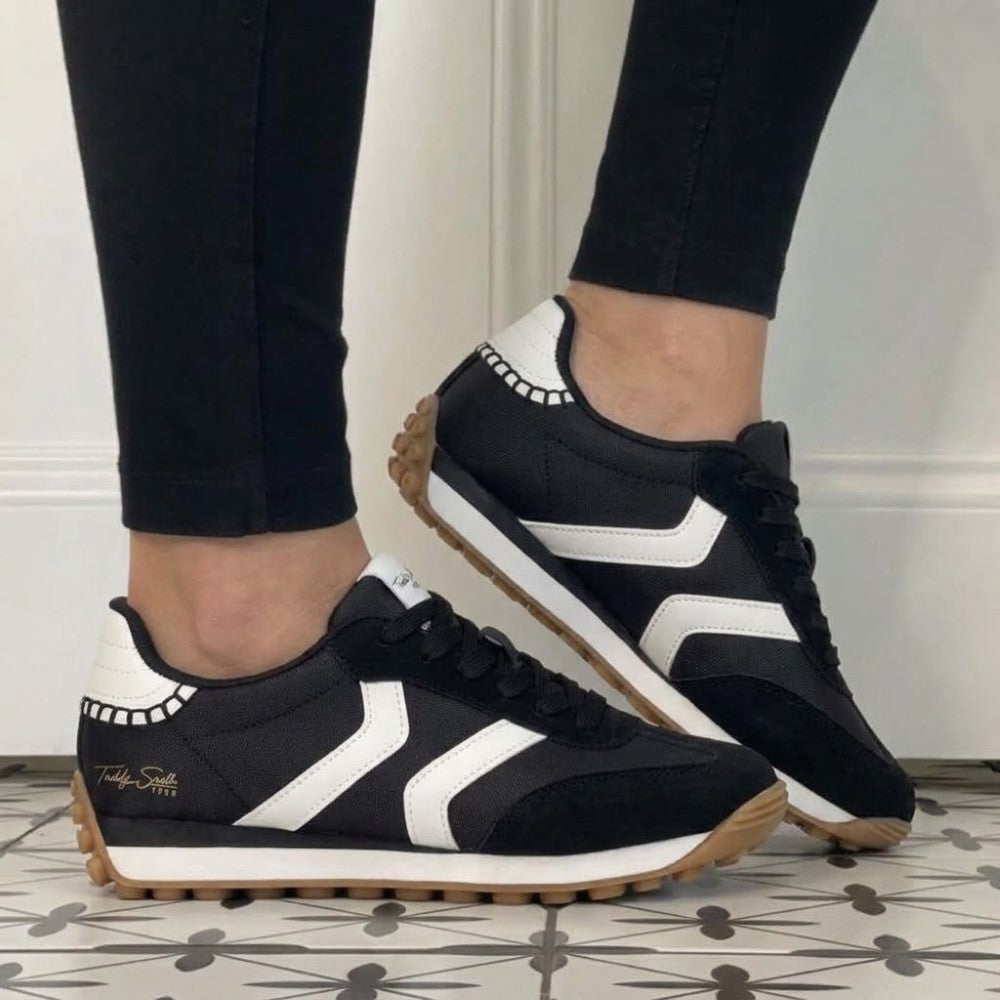 A person wearing Teddy Smith black sneakers with bold white side stripes, black laces, and gum rubber soles, paired with black leggings. The shoes are shown on a patterned tile floor against a white panelled wall.