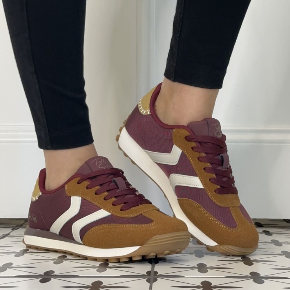 A pair of Teddy Smith low-top sneakers shown being worn, with a person's lower legs visible in black leggings. The shoes feature a burgundy upper, tan suede overlays, white side stripes, and a white midsole with a gum outsole.