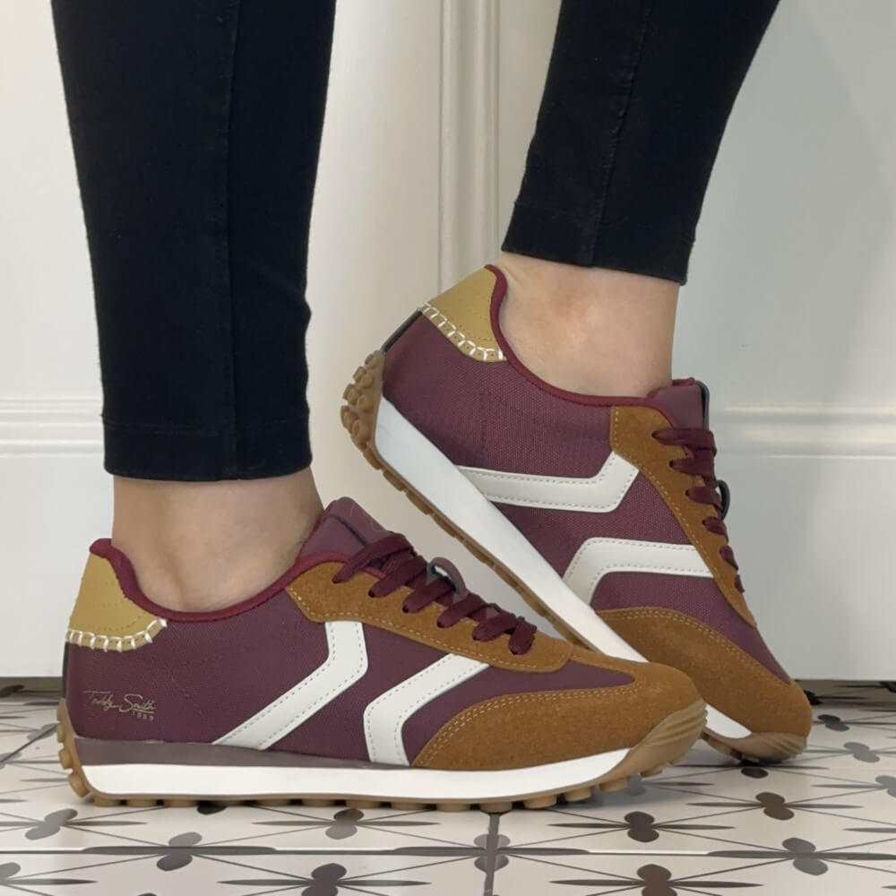 A pair of Teddy Smith low-top sneakers shown being worn, with a person's lower legs visible in black leggings. The shoes feature a burgundy upper, tan suede overlays, white side stripes, and a white midsole with a gum outsole.