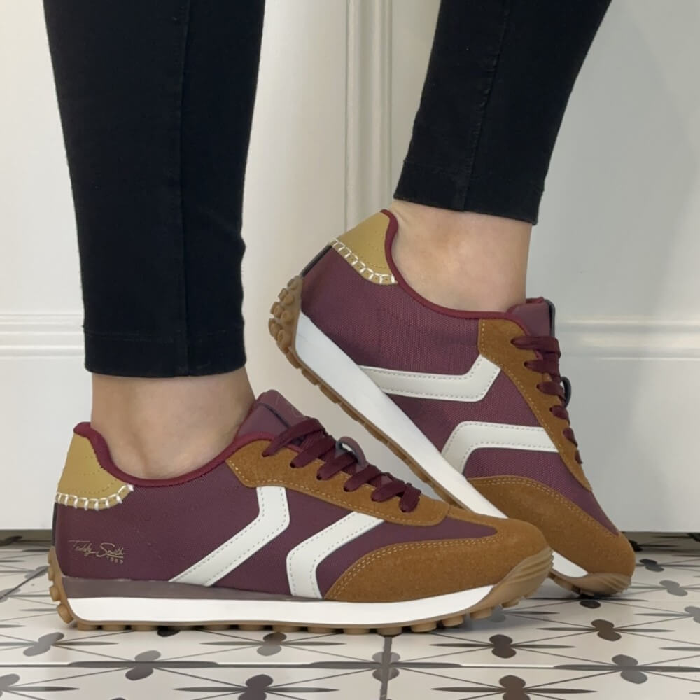 A pair of Teddy Smith low-top sneakers shown being worn, with a person's lower legs visible in black leggings. The shoes feature a burgundy upper, tan suede overlays, white side stripes, and a white midsole with a gum outsole.
