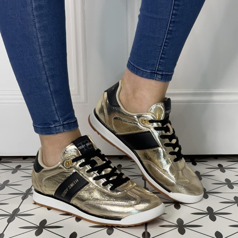 Gold and black sneakers worn with blue jeans on a patterned floor.