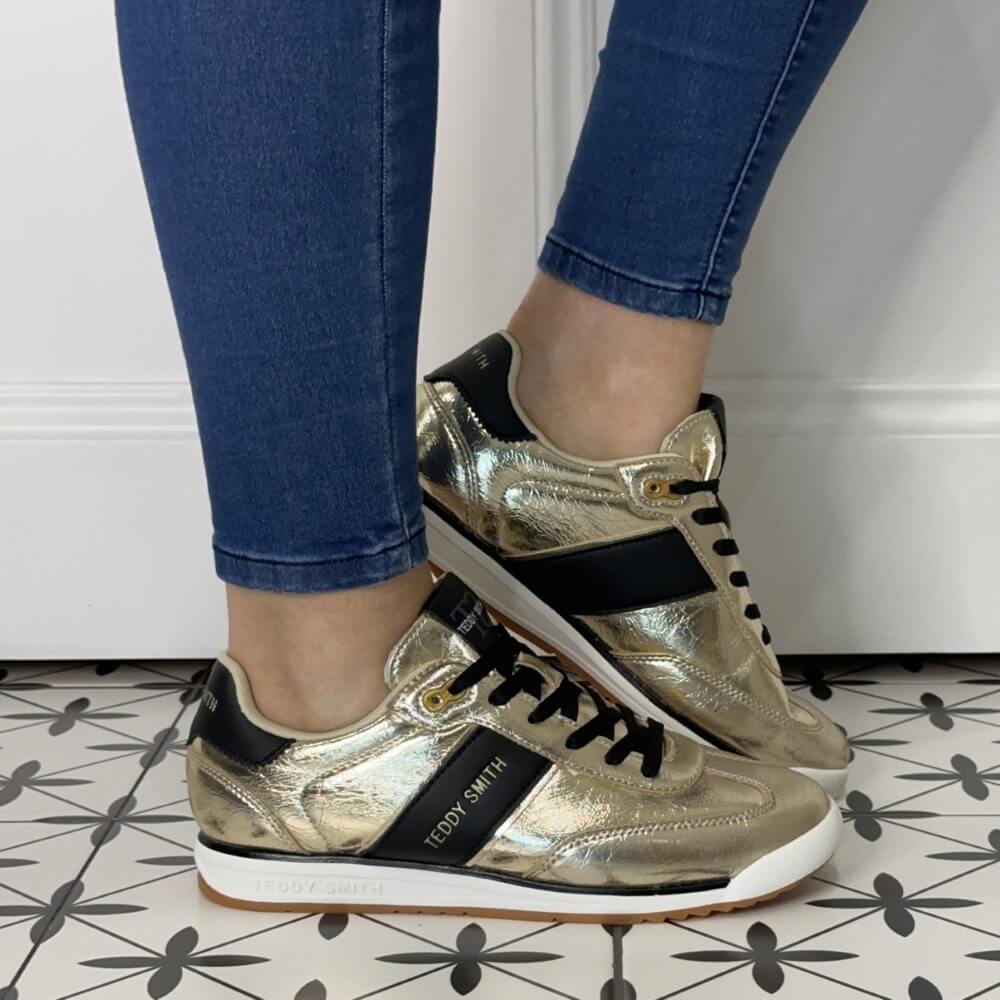 Gold sneakers with black accents worn with blue jeans on a patterned floor.