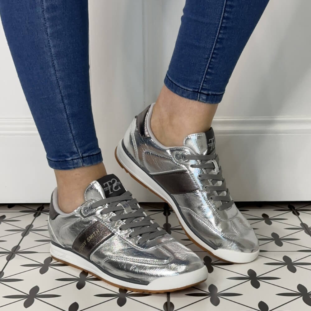 Silver sneakers worn with blue jeans on a patterned floor.
