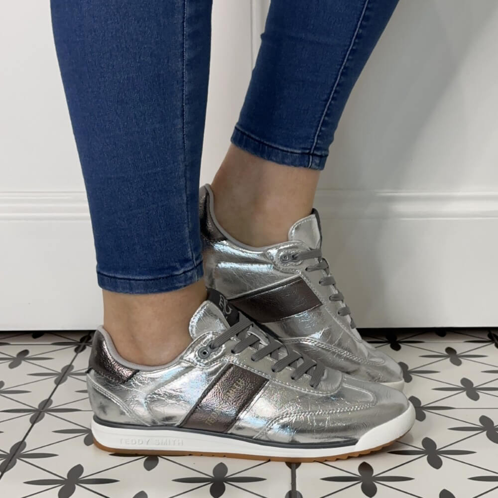 Silver sneakers worn with blue jeans on a patterned floor.