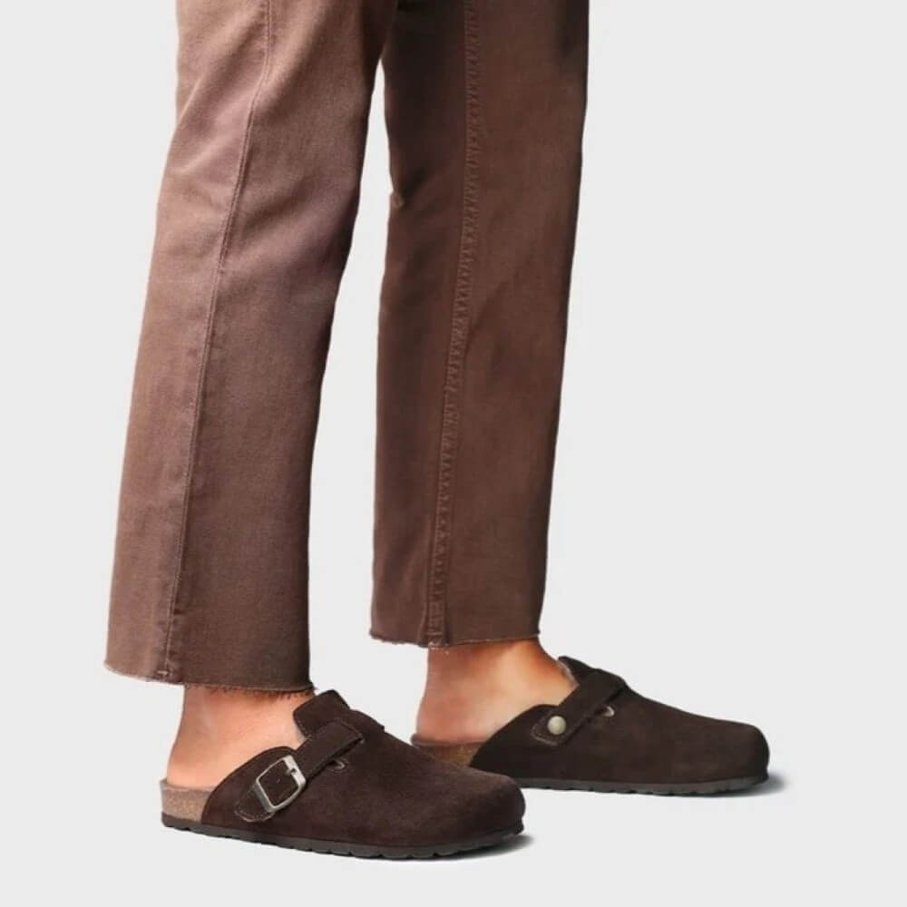 A person wearing brown suede clog-style mules with metal buckle straps, shown from the calves down against a light gray studio background.