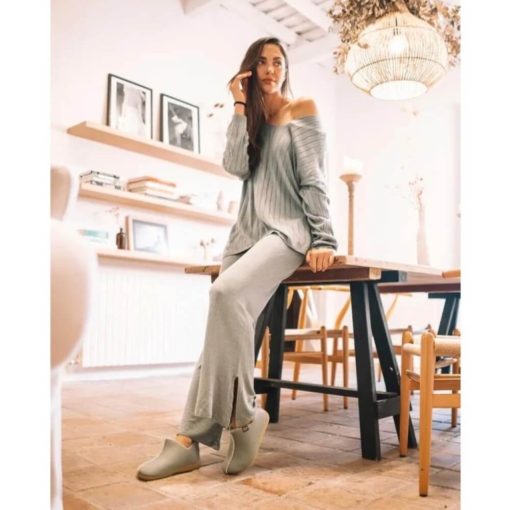 A woman wearing Toni Pons Mint Slippers, a light grey ribbed knit lounge set consisting of an off-the-shoulder long-sleeve top and matching wide-leg pants, sitting on a wooden table in a bright, modern room.
