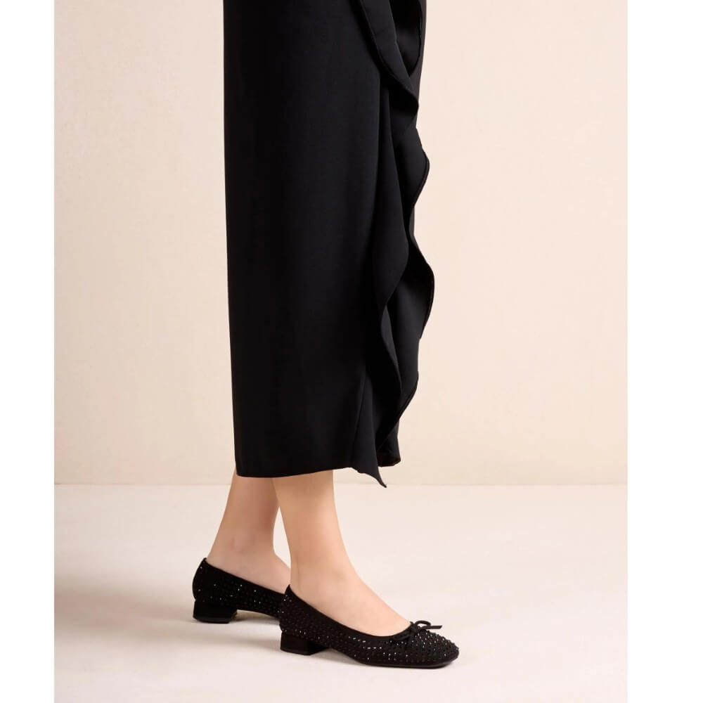 Unisa Black flat shoes with small bow details and subtle rhinestone embellishments, shown on a model wearing black draped trousers against a light beige studio background.
