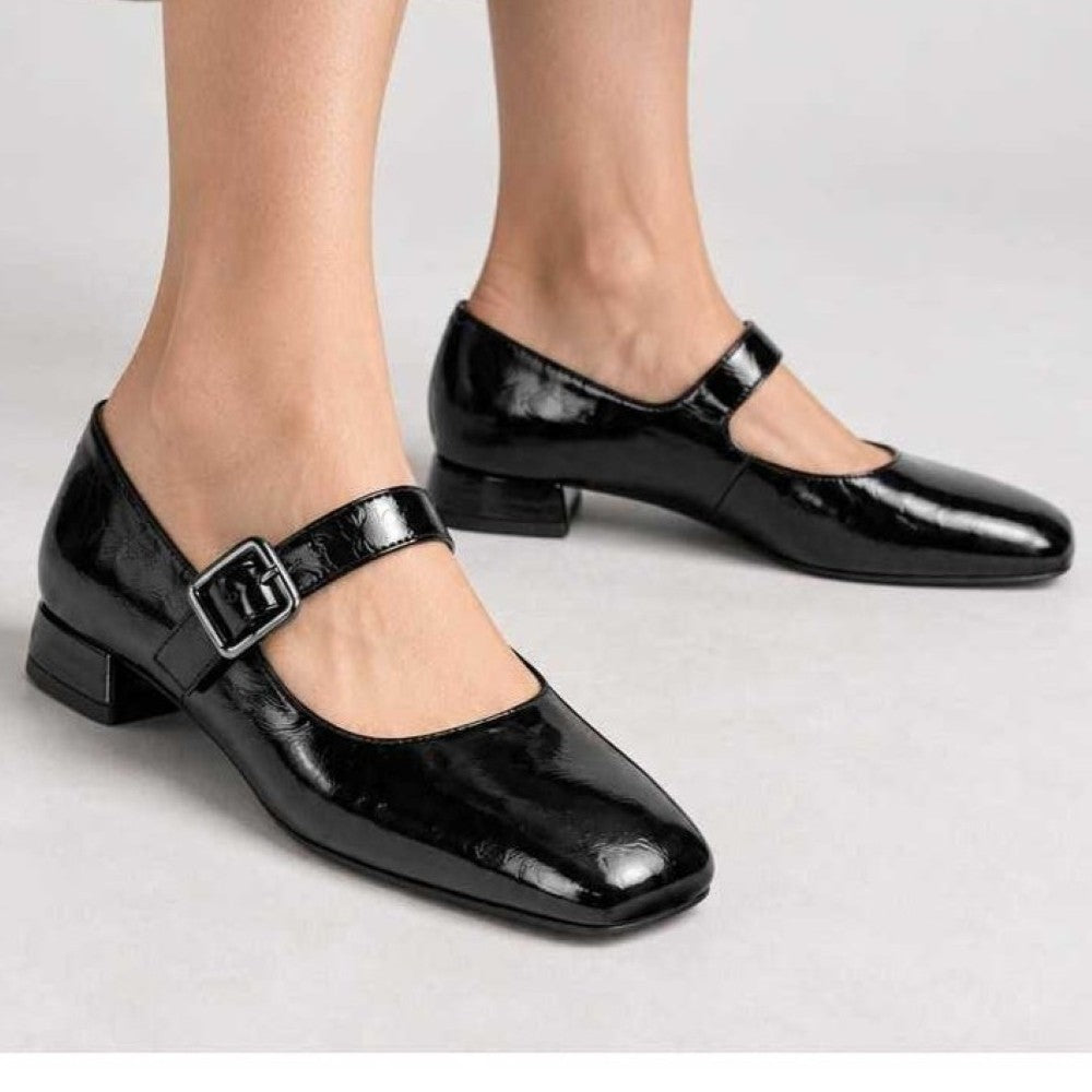 A pair of Unisa black patent leather Mary Jane shoes with low block heels and square toes, shown on a model's feet against a light grey studio background.