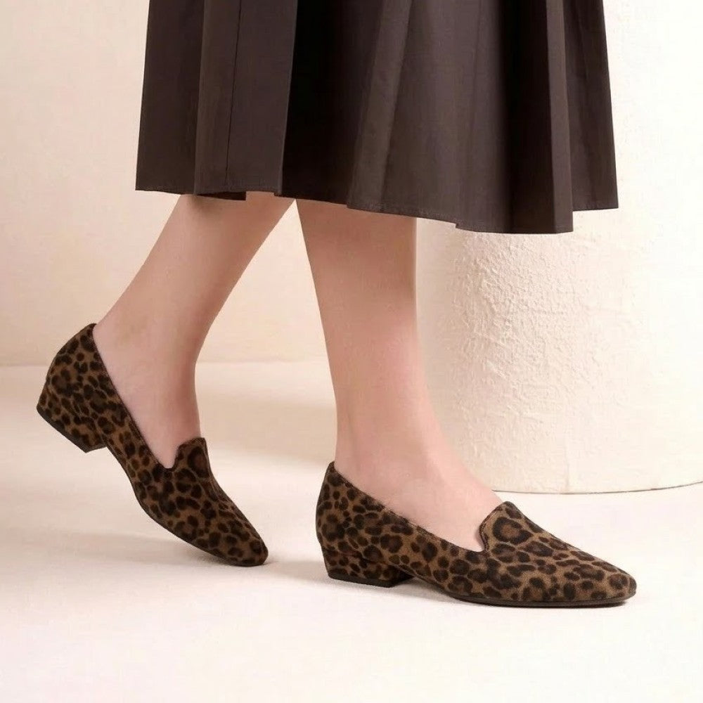 A person wearing Unisa leopard-print slip-on loafers with low block heels, paired with a dark brown pleated midi skirt, standing against a light beige textured wall.