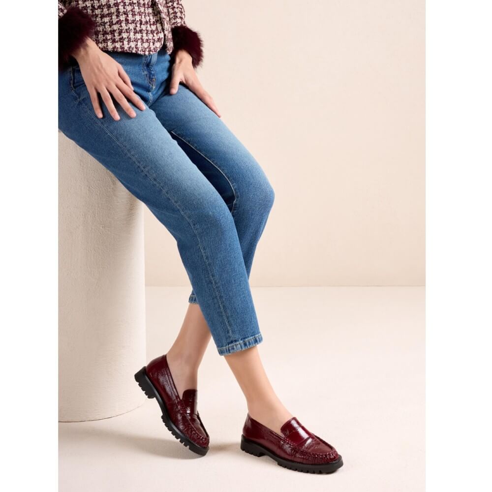 A model wearing blue cropped jeans and Unisa glossy burgundy leather penny loafers, sitting on a beige cylindrical pedestal against a neutral beige background.
