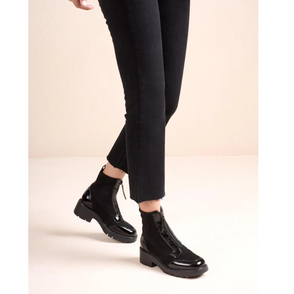 A person wearing Unisa black patent leather ankle boots with front zippers and chunky lug soles, paired with black cropped jeans, against a light beige studio background.