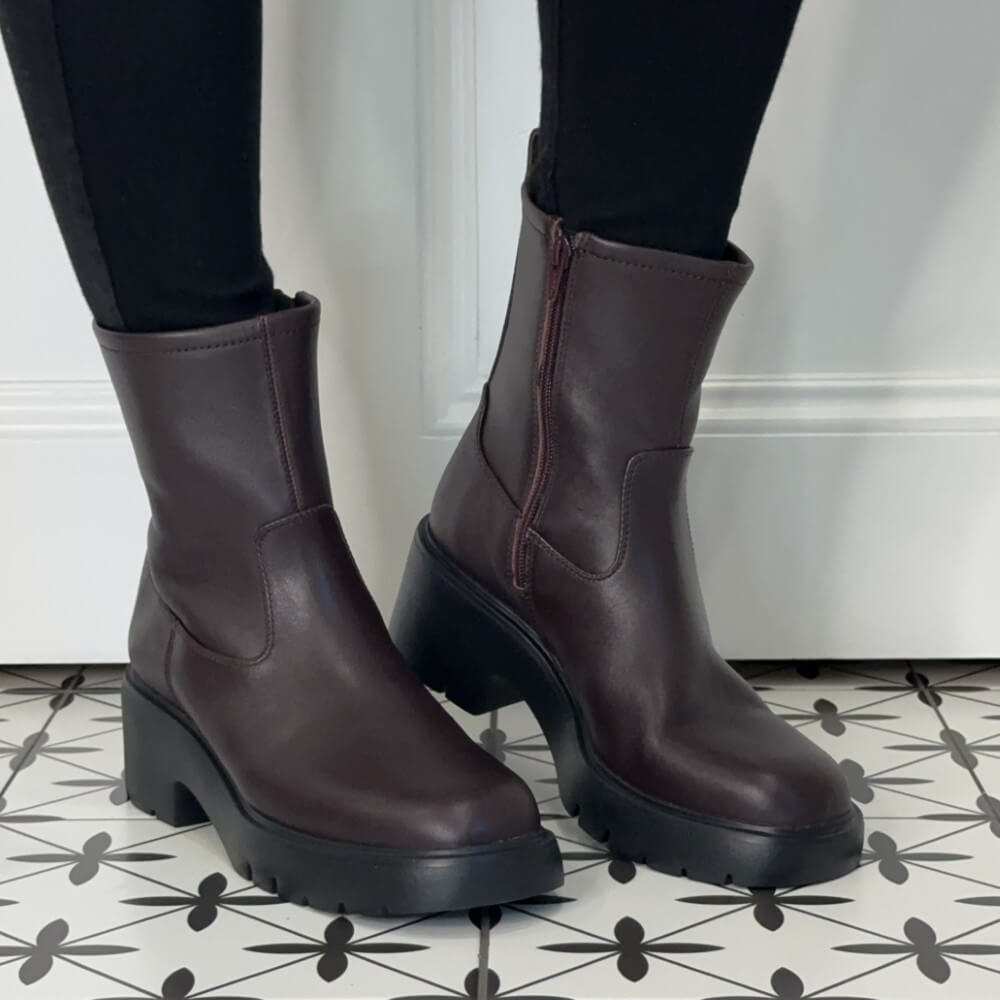 Unisa wine leather ankle boots with a side zipper and chunky black platform soles, shown on a person wearing black leggings standing on a black-and-white patterned tile floor.