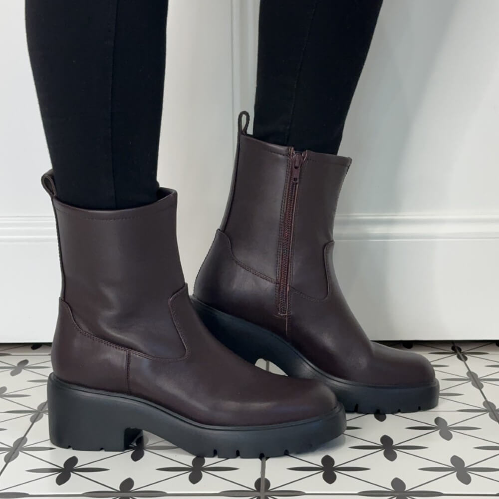 Unisa wine leather ankle boots with a side zipper and chunky black platform soles, shown on a person wearing black leggings standing on a black-and-white patterned tile floor.