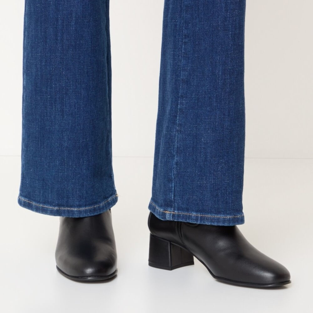 Close-up of a person wearing dark blue flared jeans and Unisa black leather ankle boots with a block heel, shown against a plain light background.