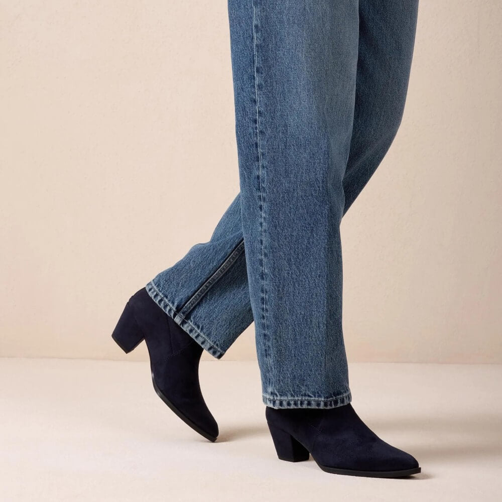 A person wearing blue jeans and Unisa Black pointed-toe heeled ankle boots, shown from the knees down against a plain beige studio background.