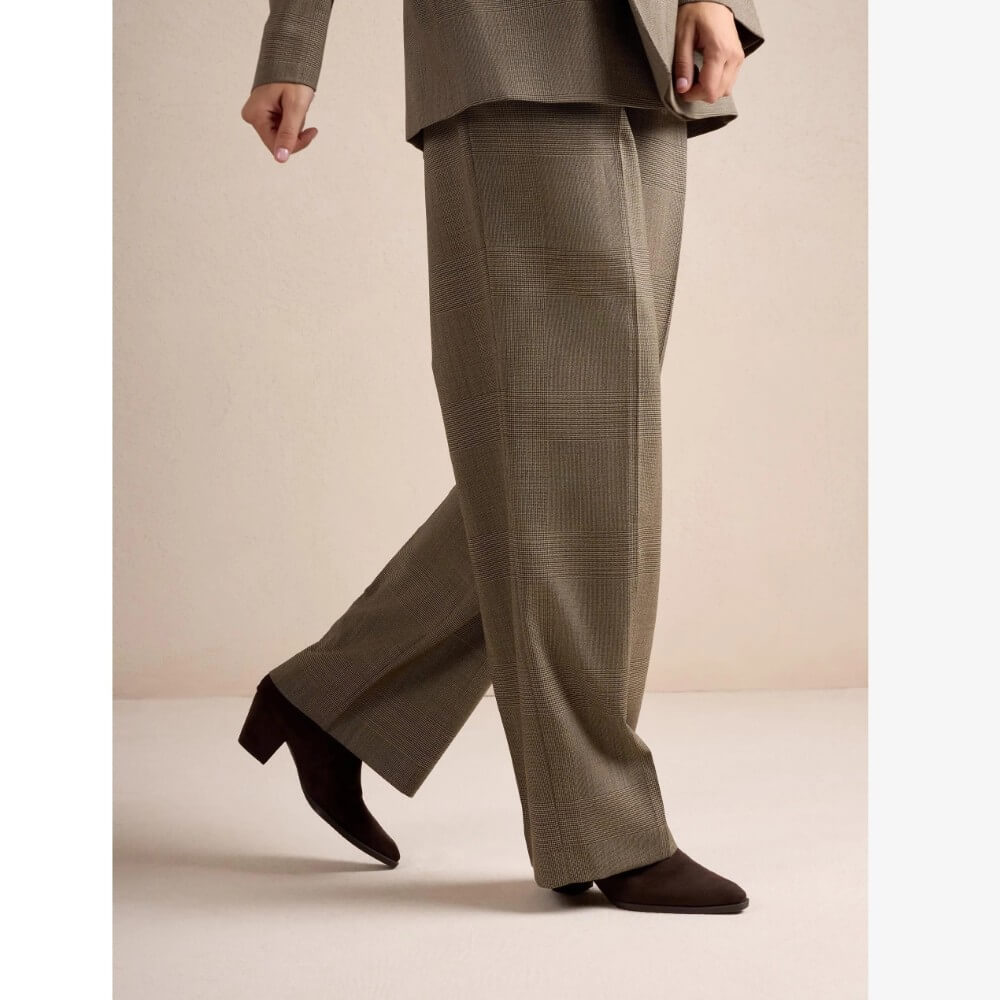 A person wearing wide-leg taupe trousers with a subtle plaid pattern, styled with a matching blazer and Unisa dark brown pointed-toe ankle boots, shown from the waist down against a neutral beige studio background.