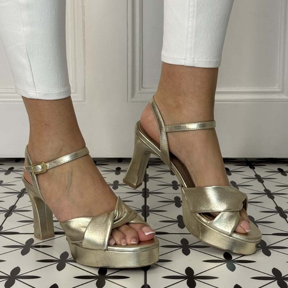 Gold high-heeled sandals worn with white pants on a patterned floor.